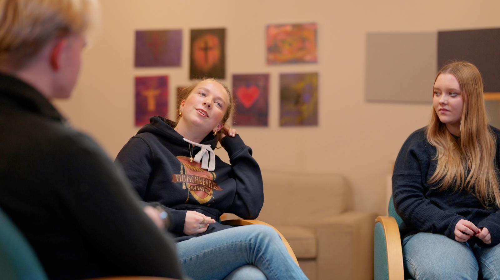 To unge jenter sitter og diskuterer, mens en gutt sitter i forgrunnen og hører på